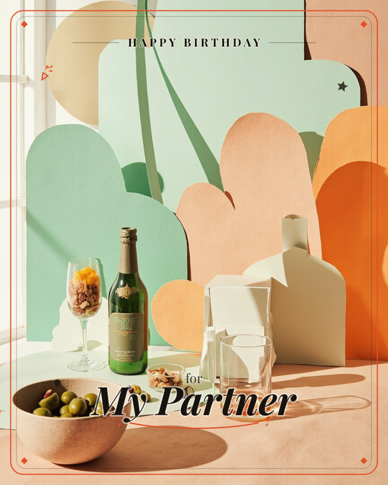 A styled still life shows a bottle of sparkling wine, a bowl of green olives, a glass of fruit-topped dessert and a low tumbler of something amber, all…, a birthday e-card design