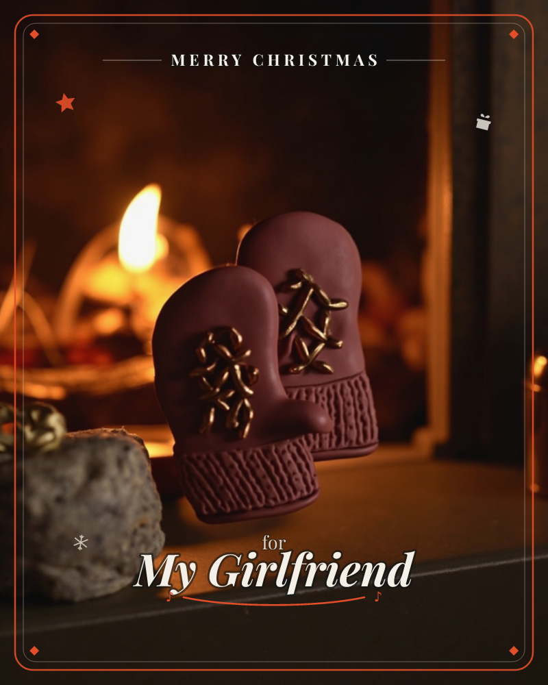 A pair of small ceramic mittens with knitted-texture cuffs and gold thread lacing sit on a dark mantel, glowing warm maroon in candlelight, a christmas e-card design