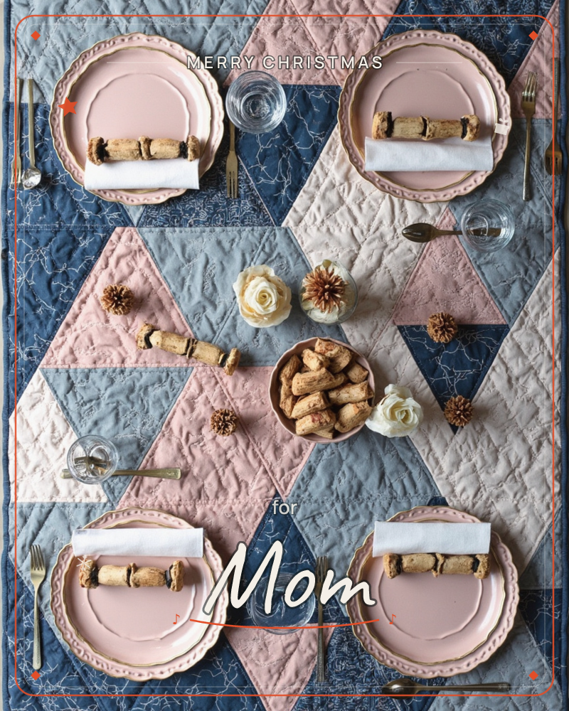 An overhead view of a festive table set on a patchwork quilt of navy, blush pink and cream triangles, with pink-rimmed plates, folded white napkins…, a christmas e-card design