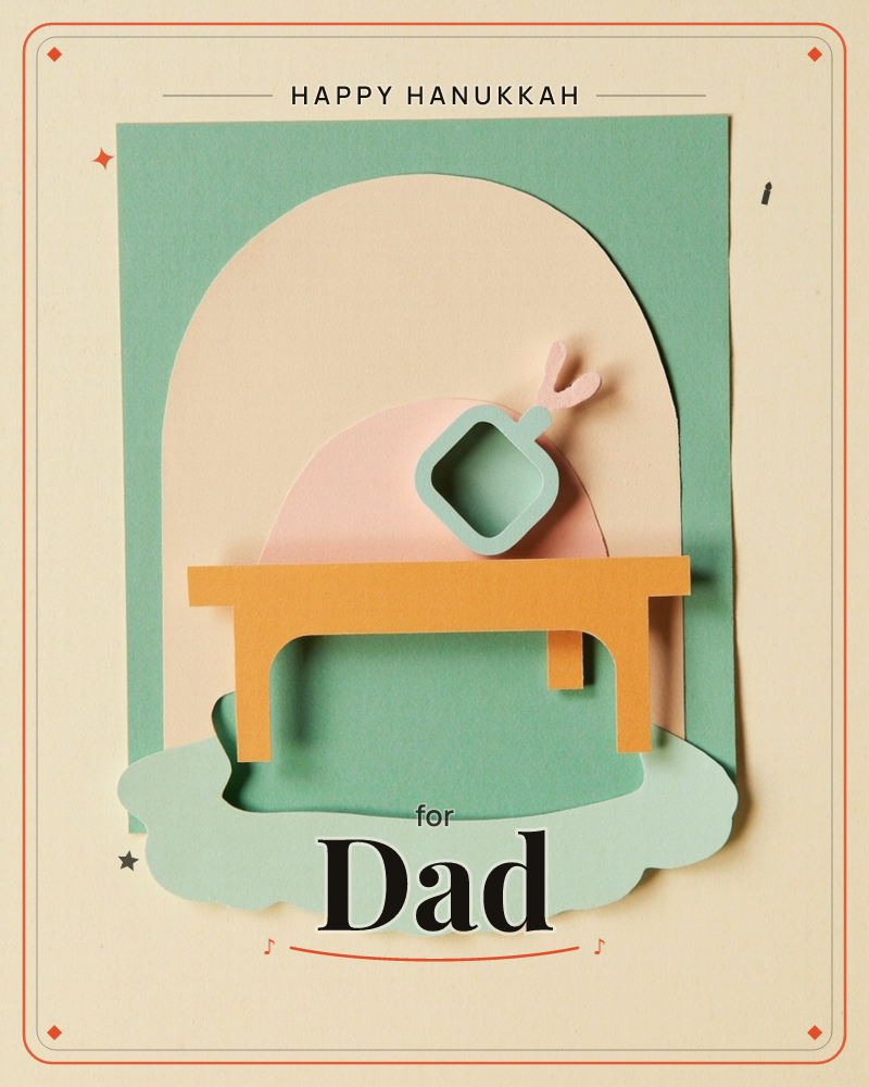 A paper-cut illustration shows a teal frying pan tilted on an orange table, tossing something into the air above a pale pink archway, a hanukkah e-card design