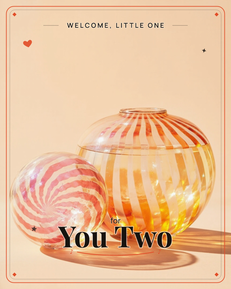Two blown-glass forms sit against a warm peach backdrop: a small striped sphere in candy-pink swirls beside a larger amber vase ribbed with orange and…, a new baby e-card design