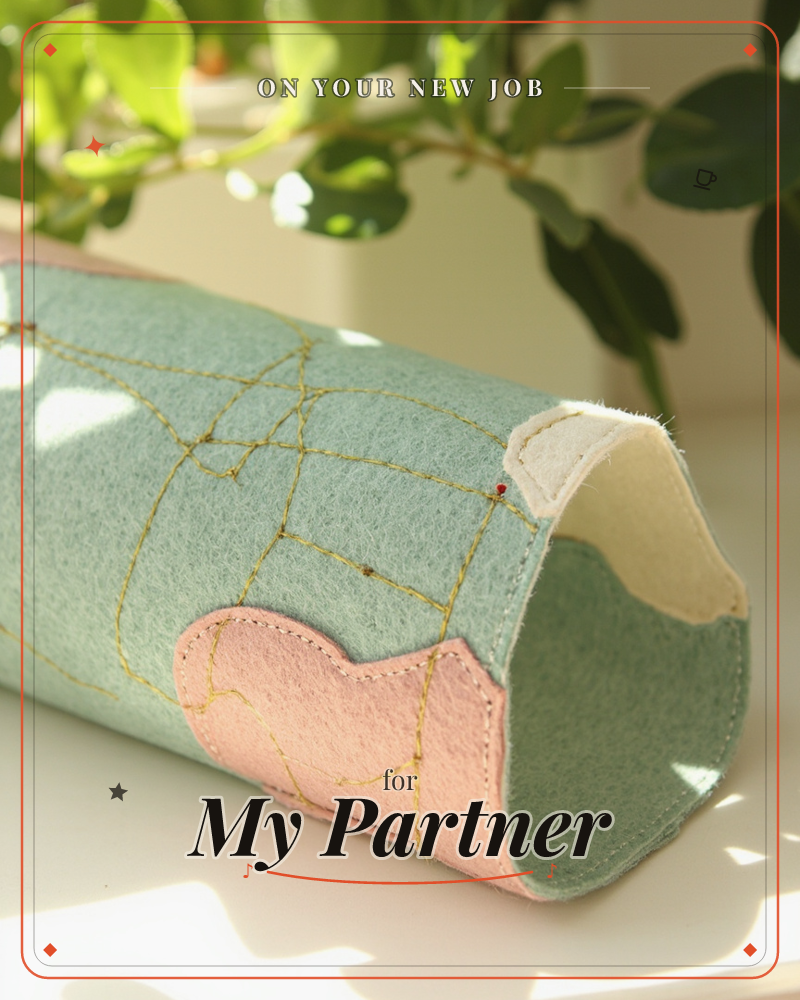 A soft felt cylinder, stitched in sage green and dusty pink with wandering gold thread lines like a hand-drawn map, sits on a sunlit windowsill beneath…, a new job e-card design