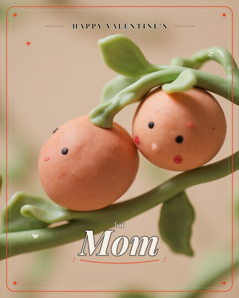 A close-up of two round clay fruits with tiny painted faces, nestled together on a green sculpted vine with curling leaves, a valentine e-card design